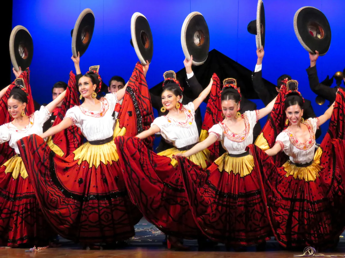 Dancers of the Ballet Folklórico de México de Amalia Hernández company on stage in wide red, black and gold Jalisco-style skirts, arms raised holding wide-brimmed sombreros, mid-performance