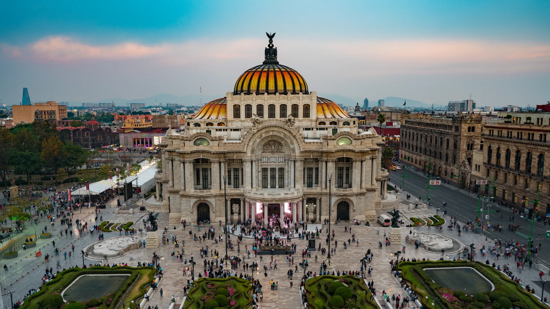 The Palacio de Bellas Artes at dusk, a white and beige marble Art Deco and Art Nouveau palace topped with an orange-and-cream tiled dome, seen from above with its plaza and formal gardens in front, Mexico City