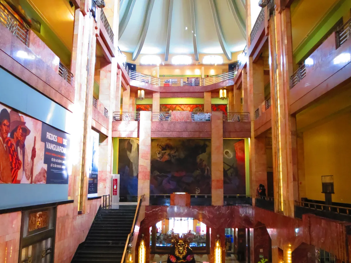 The Art Deco vestibule inside the Palacio de Bellas Artes, showing rose-toned marble columns, a curved skylight and murals on the upper gallery walls