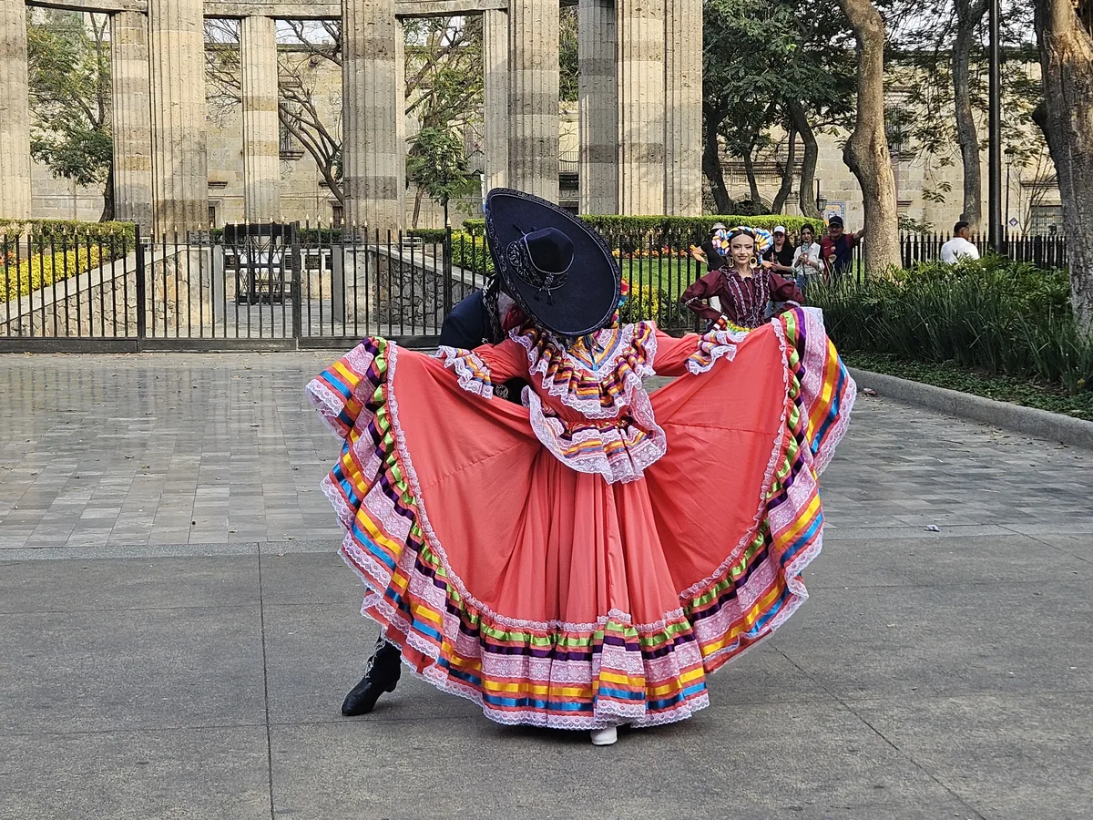 A dancer in a wide, colourfully trimmed coral-pink Jalisco-style skirt performing the Jarabe Tapatío outdoors in Guadalajara, illustrating the regional costume style — not a photo of the Bellas Artes show itself